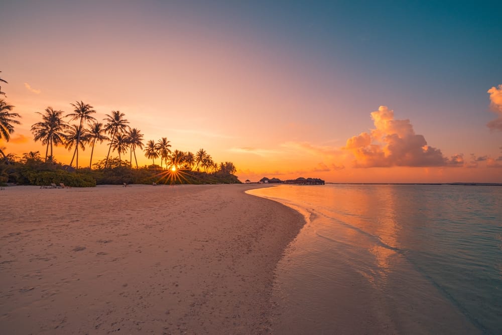 Tropisk strand med farverig himmel ved solnedgang – gyldent sand, solstråler over havet og afslappende ferieatmosfære.