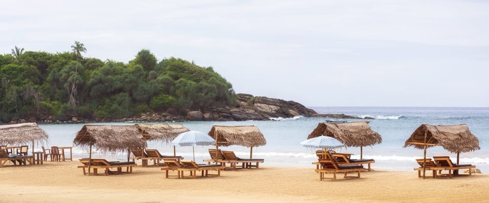 En fredelig strand i Sri Lanka med stråparasoller og blødt gyldent sand. Det krystalklare hav og de tropiske omgivelser skaber en idyllisk atmosfære – perfekt til afslapning, solbadning og ferieoplevelser ved kysten.