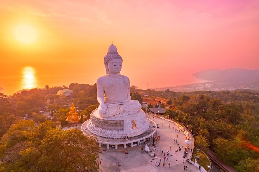 Luftfoto i topview af den imponerende Big Buddha-statue i Phuket ved solnedgang. Den majestætiske hvide Buddha rejser sig over bjergryggen med panoramaudsigt over øen, omgivet af gyldne himmelnuancer – et spirituelt vartegn og populært rejsemål i Thailand.