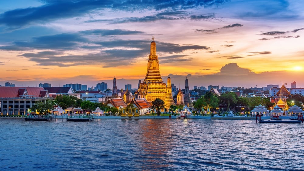 Luftfoto af Wat Arun-templet (Wat Arun Ratchawararam) i Bangkok ved solnedgang. Det berømte tempel, også kaldet Daggryets Tempel, står smukt oplyst med gyldne farver, der spejler sig i Chao Phraya-floden – et ikonisk vartegn og populært rejsemål i Thailand.