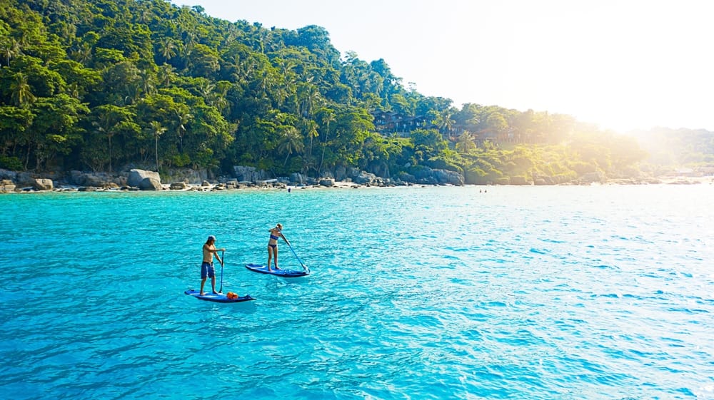 Par på paddleboard i havet om sommeren – droneudsigt over klart vand og udendørs vandsportoplevelse. » Tembo Travel Par på paddleboard i havet om sommeren – droneudsigt over klart vand og udendørs vandsportoplevelse.