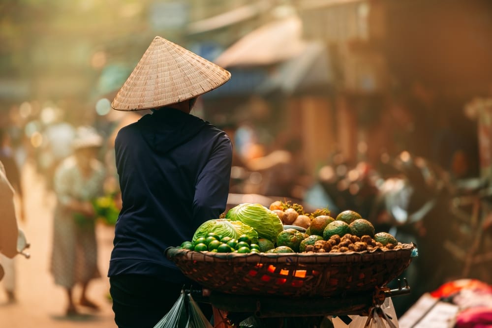 En lokal kvinde går gennem de stemningsfulde gader i Hoi An med kurve fyldt med frugt på vej til markedet – et autentisk glimt af hverdagslivet.