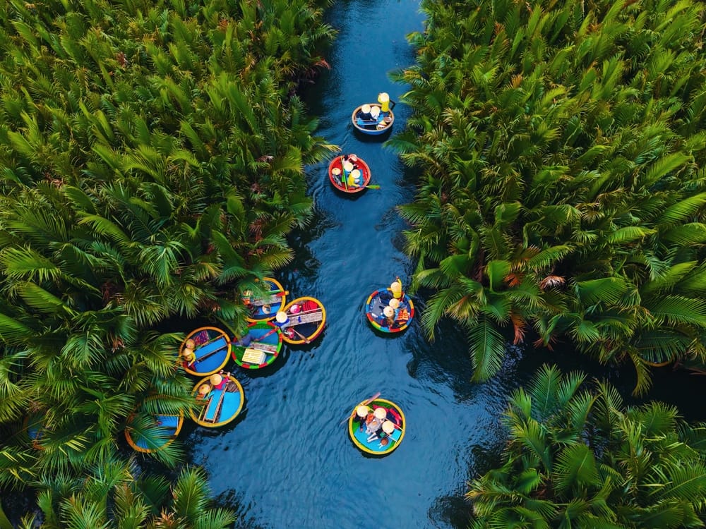 Luftfoto af traditionelle kokos- og bambusbåde, der glider stille gennem smaragdgrønne kanaler omgivet af frodig vegetation.