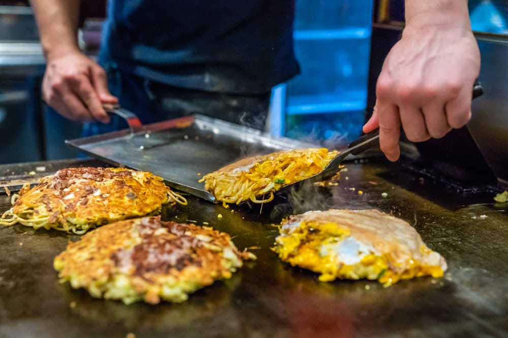 Japansk kok tilbereder Hiroshima-style okonomiyaki på varm jernplade.