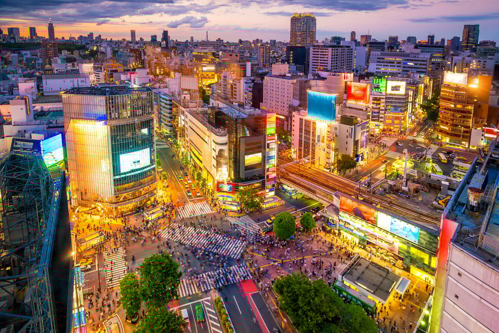 Topudsigt over Shibuya Crossing i Tokyo, Japan, ved skumringstid, hvor tusindvis af fodgængere krydser under byens lys og skærme