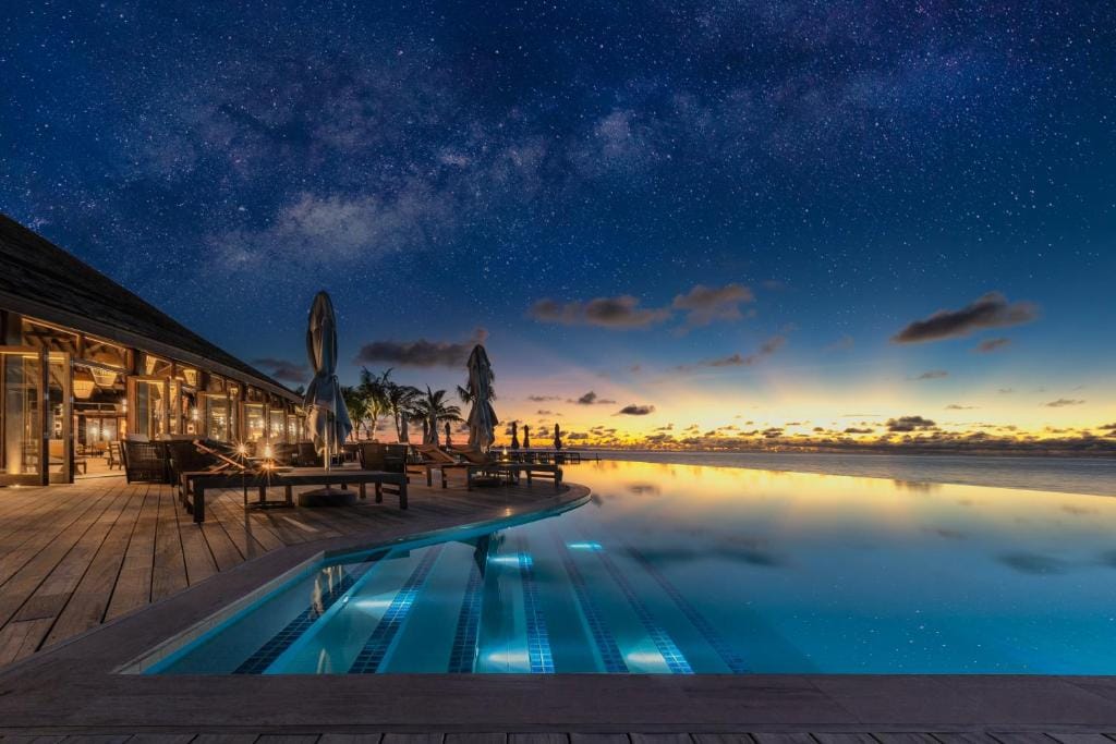 Infinity-pool om aftenen med stjernehimmel og oplyst restaurant på Jawakara Maldiverne.