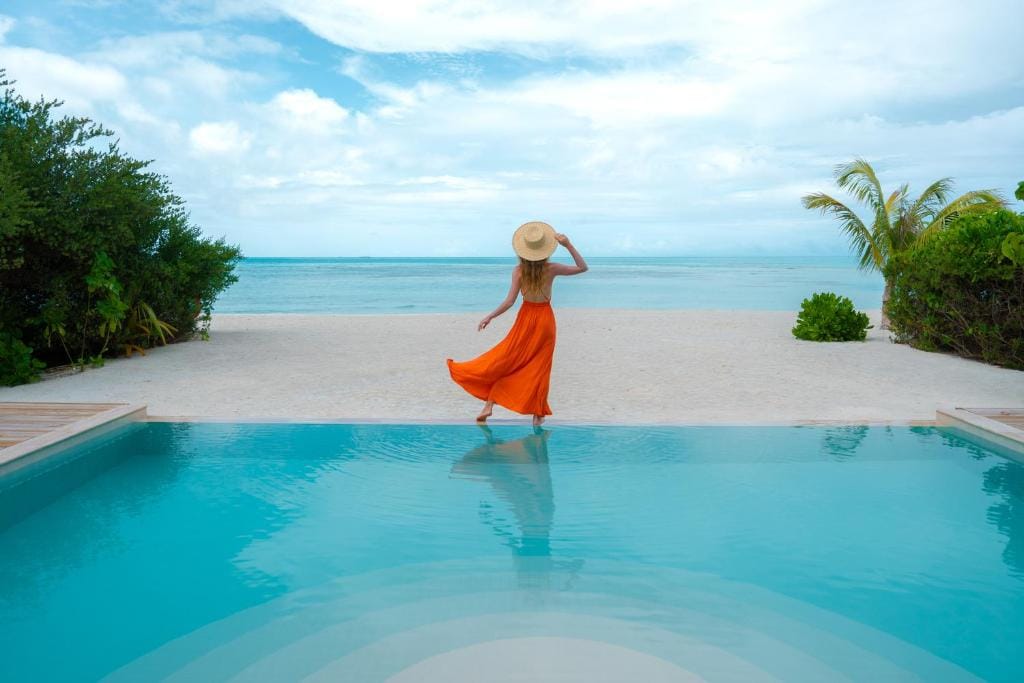 Kvinde i orange kjole og solhat står ved pool med udsigt til strand og turkis hav på Jawakara Islands Maldiverne.