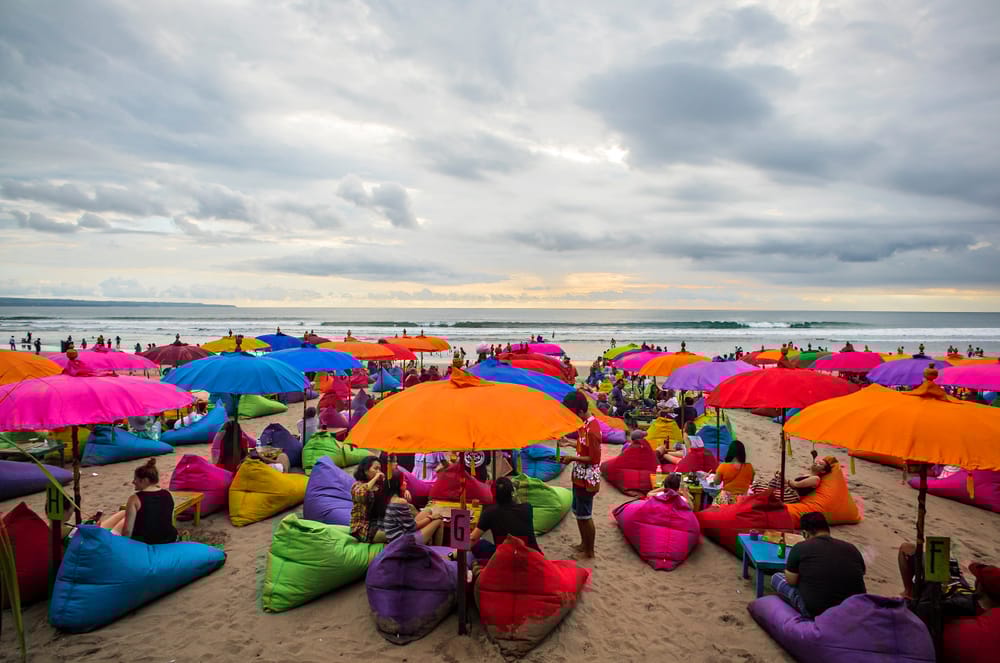 Turist på Seminyak Beach, Bali, Indonesien, med farverige parasoller og solnedgang over havet.