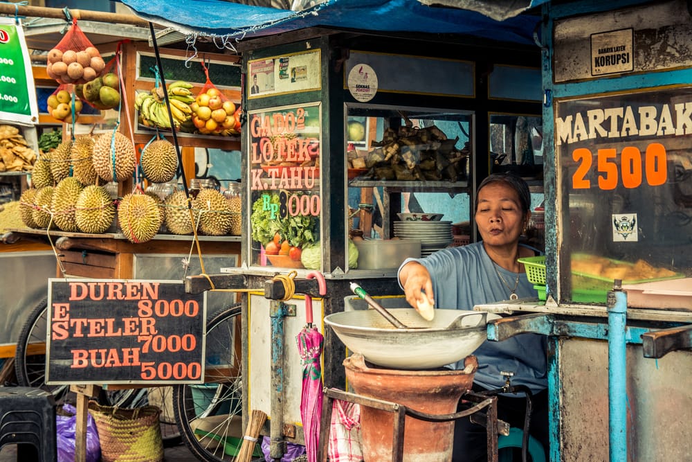 Lokal street food-sælger på scooter på Bali, Indonesien, omgivet af byliv og farver.