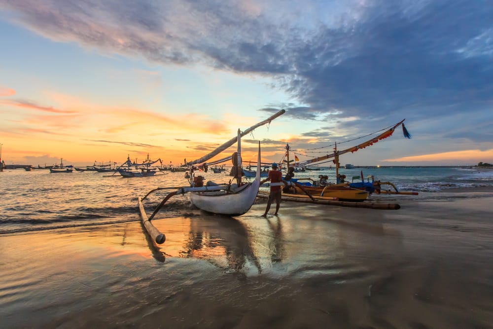 Solnedgang ved Jimbaran Beach, Bali, Indonesien, med palmer, hav og strandrestauranter i aftenskær.