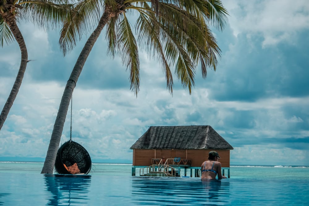 Kvinde i infinity pool med udsigt til palmer og en water villa på Maldiverne.