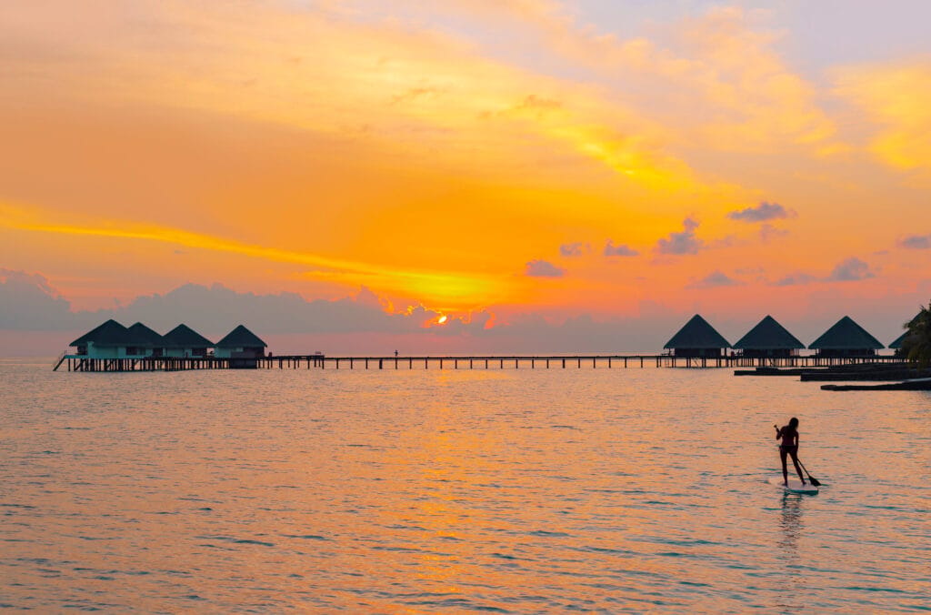Person på paddleboard ved solnedgang foran overwater villas på Finolhu Maldiverne.