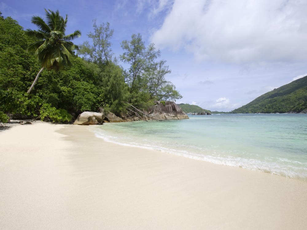 Strand med klart, lavvandet hav ved Port Launay Marine National Park på Mahé, Seychellerne, omgivet af grønne bjerge.