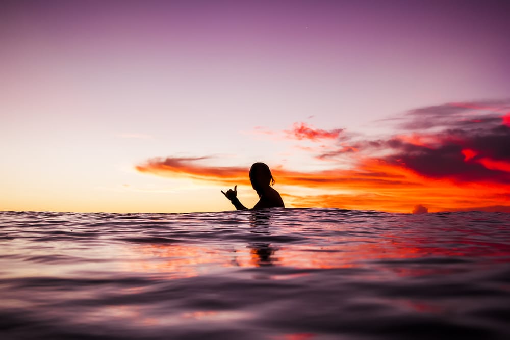 Surfer ved solnedgang på stranden i Bali, Indonesien, med orange himmel og rolige bølger.