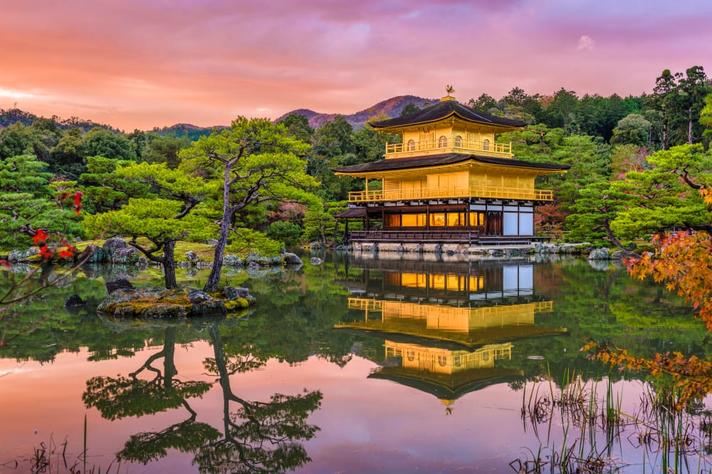 Kinkaku-ji, Den Gyldne Pavillon i Kyoto, der spejler sig i en rolig sø omgivet af grønne træer og farverige efterårsblade ved solnedgang.