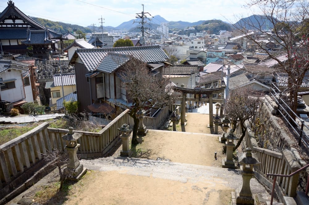 Stentrappe i Onomichi med stenlanterner, torii-port og udsigt over byens tage og bjerge i Hiroshima-præfekturet.