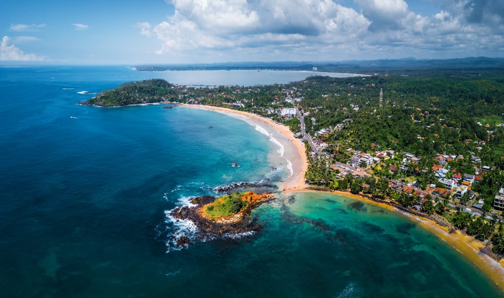 Panoramisk luftfoto af Mirissa i Sri Lanka med sandstrand, palmer, bebyggelse og Det Indiske Ocean.