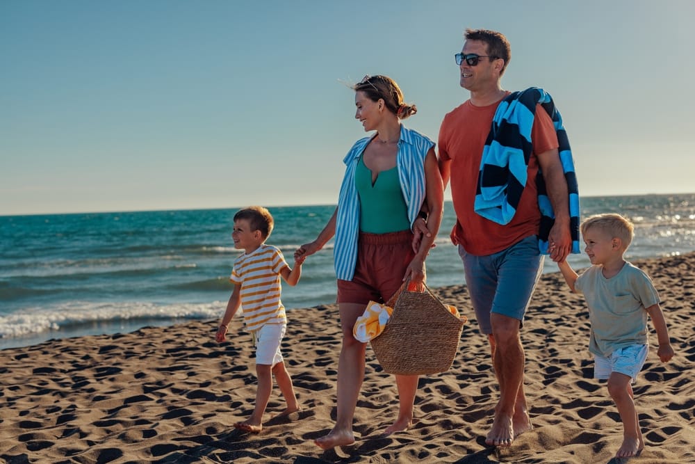 Familie med to voksne og to børn går hånd i hånd langs sandstranden ved havet.