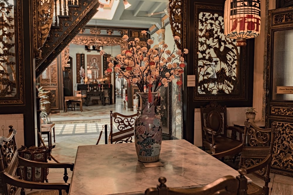 Traditionelt Peranakan-interiør med udskårne træpaneler, antikke møbler og vase med blomster.