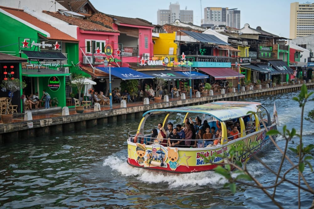 Farverig turistbåd på Malacca River med restauranter og farverige bygninger langs floden.