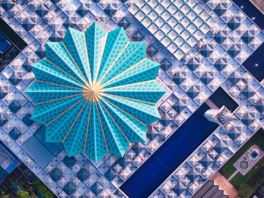 Luftfoto af Masjid Negara i Kuala Lumpur med den karakteristiske turkisblå stjerneformede kuppel og geometrisk tagdesign.