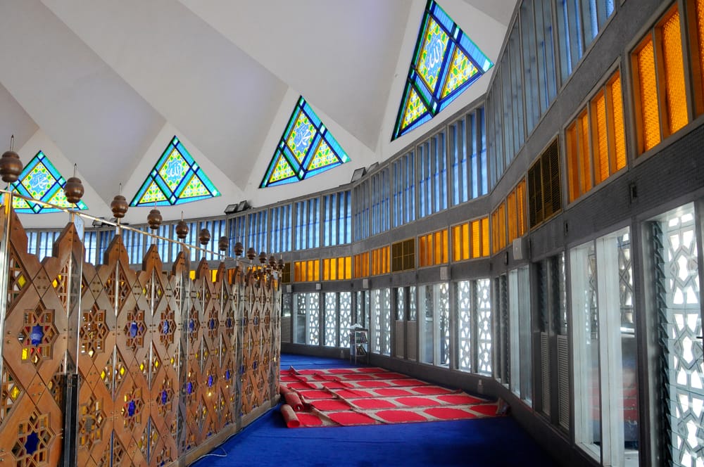 Indvendigt i Masjid Negara med farverige trekantede glasvinduer, blå tæpper og moderne islamisk arkitektur.
