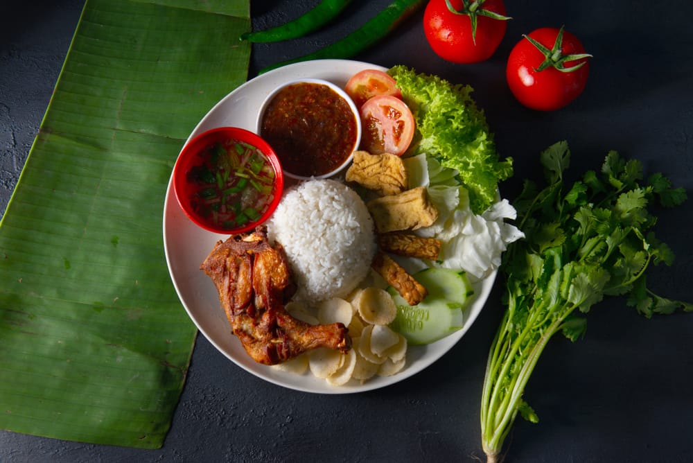 Tallerken med nasi lemak bestående af ris, sambal, stegt kylling, agurk, æg og peanuts serveret på bananblad.