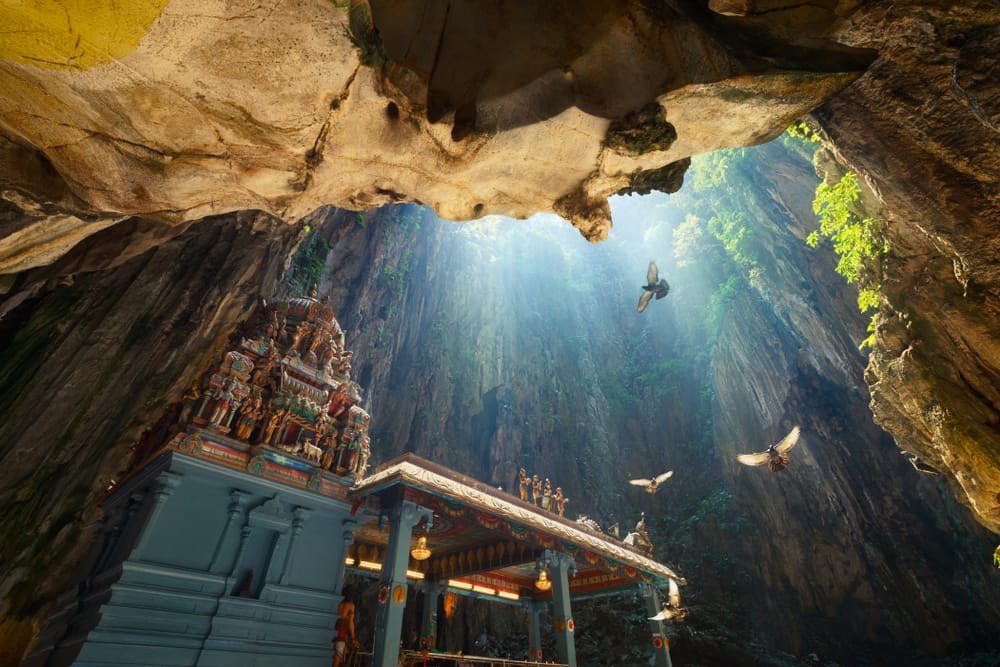 Hinduistisk tempel inde i Batu Caves med dramatisk lysindfald fra hulens åbning og stejle kalkstensvægge.