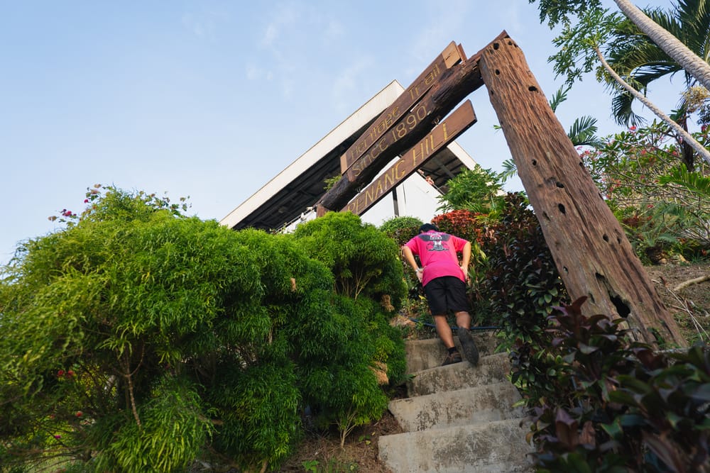Person går op ad trapper mod indgangen til Penang Hill omgivet af grøn beplantning og træskilt.