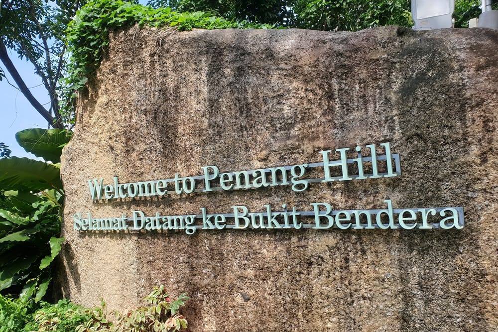 Stenskilt med teksten “Welcome to Penang Hill – Selamat Datang ke Bukit Bendera” omgivet af tropiske planter.