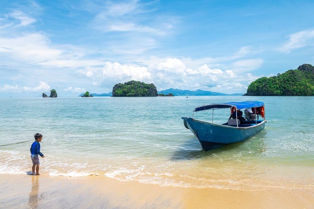 Lille dreng på sandstrand i Malaysia ser på en blå båd i det klare, turkise hav med grønne øer i baggrunden.