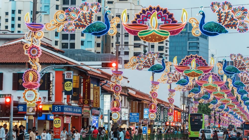 Farverige lysdekorationer over gade i Little India, Singapore, med butikker og trafik.