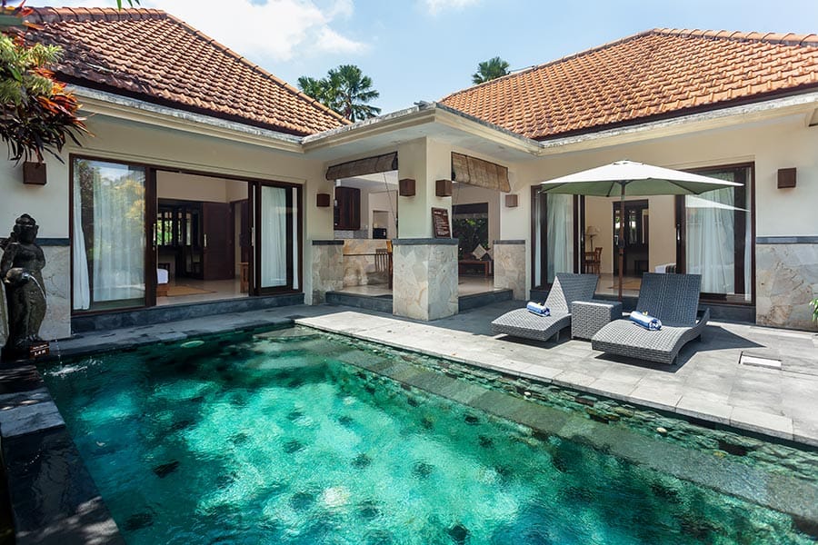 Villa courtyard with a rectangular pool, stone patio, and two lounge chairs under a large umbrella.