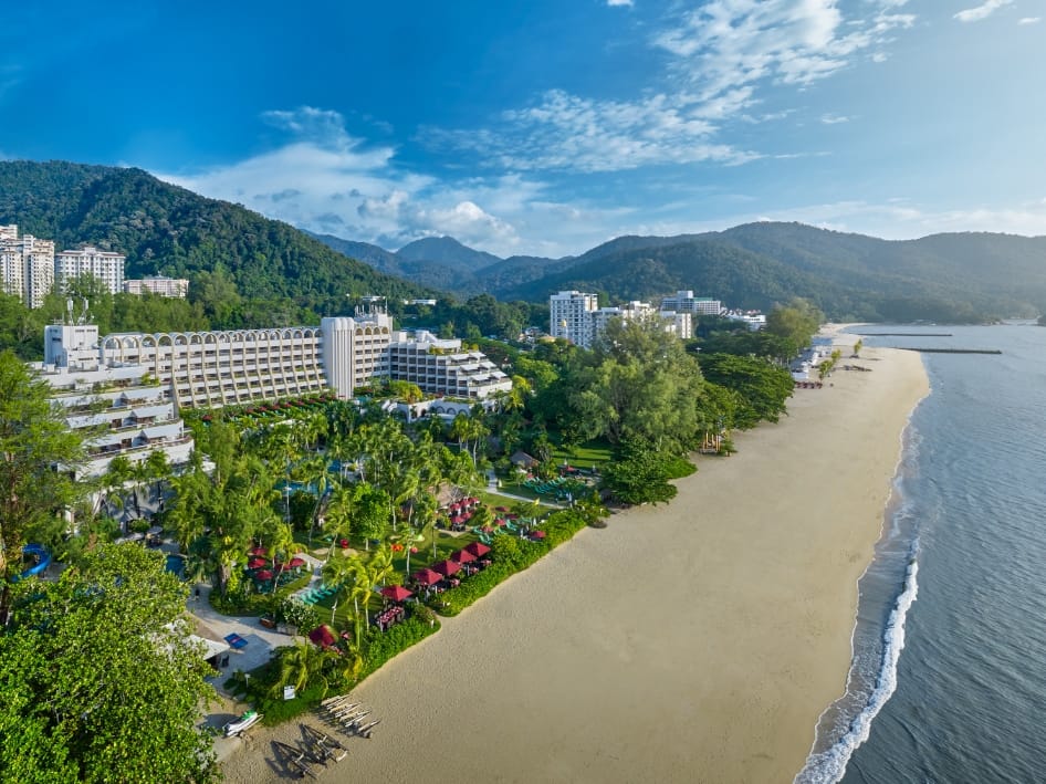 Aerial view of a tropical beach resort with white buildings, palm trees, and a long sandy shoreline along calm blue water in the bay.