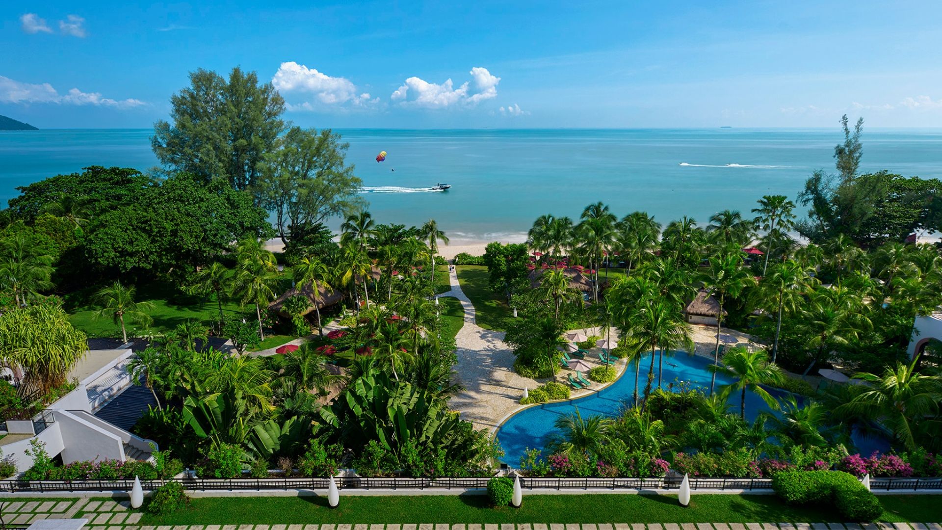 A tropical seaside resort with turquoise ocean, sandy beach, palm trees, and a winding pool area in the foreground.