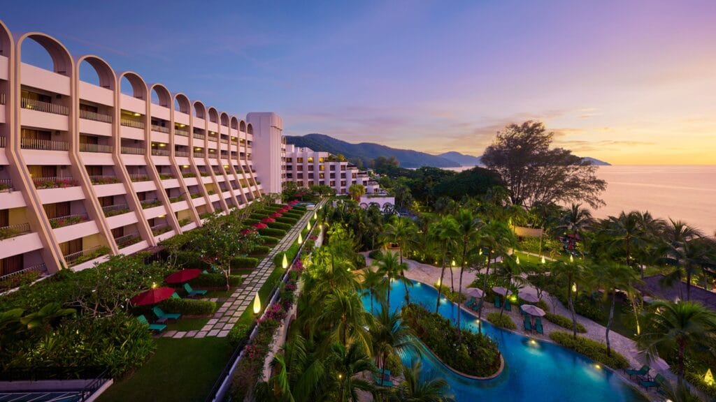 Luxury seaside resort with curved archways, lush gardens, and a winding pool at sunset overlooking the ocean.