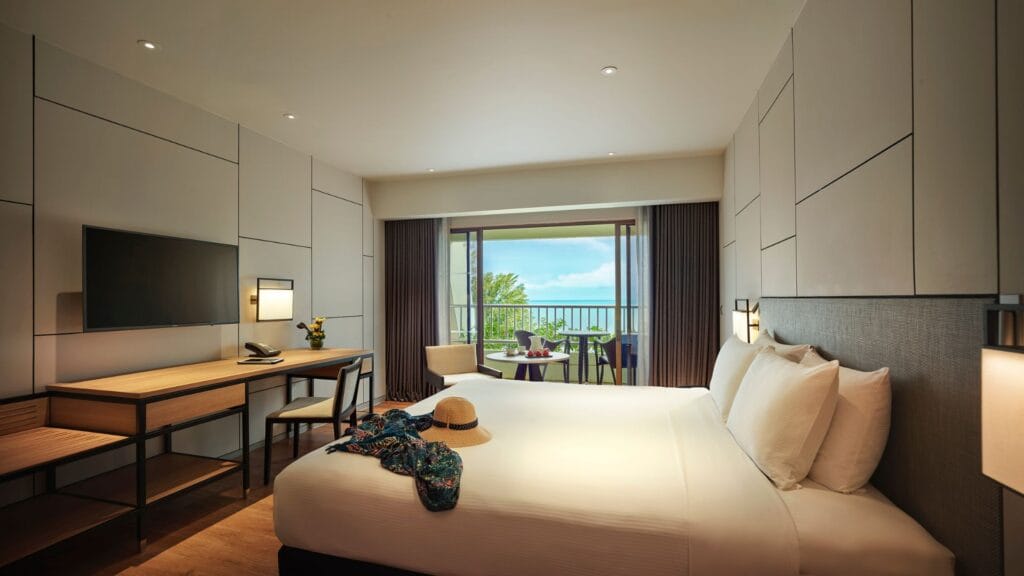 Hotel room with a large white bed, wall-paneling, a desk and chair, and a balcony with ocean view beyond sliding doors.
