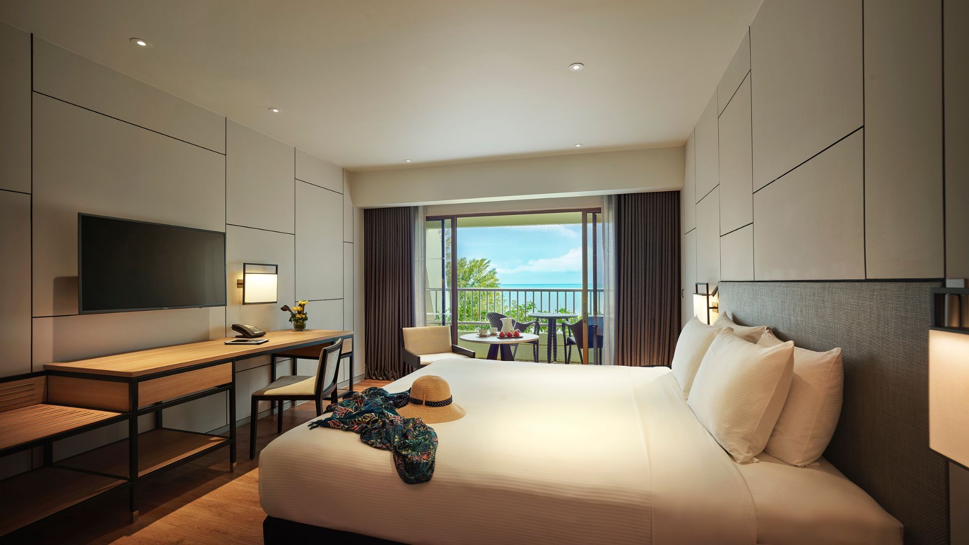 Hotel room with a large white bed, wall-paneling, a desk and chair, and a balcony with ocean view beyond sliding doors.