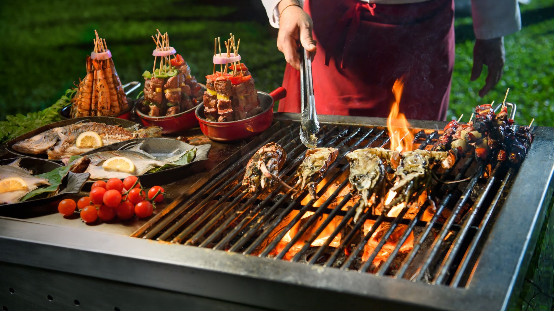 Outdoor barbecue with a variety of grilled foods: skewered meat and vegetables, whole fish with lemon, and cherry tomatoes, flames on the grill.