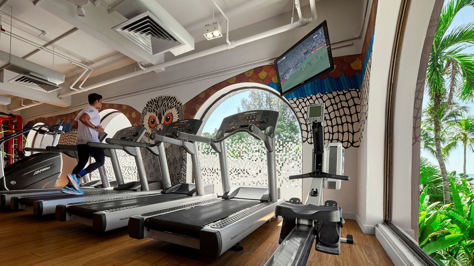 Person jogging on a row of treadmills in a bright gym with a tropical mural and large window showing palm trees outside