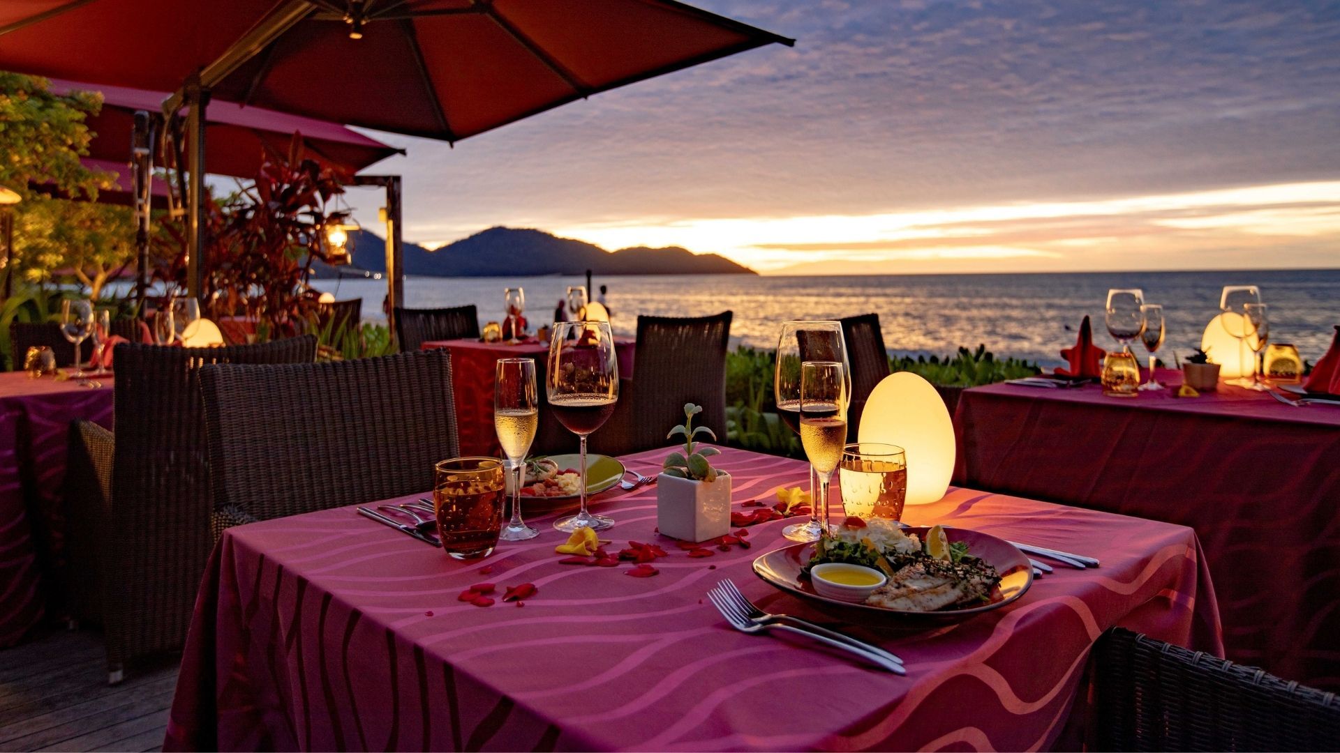 Outdoor seaside restaurant at sunset with pink tablecloths, wine glasses, and candle-like lamps overlooking the water and distant hills.