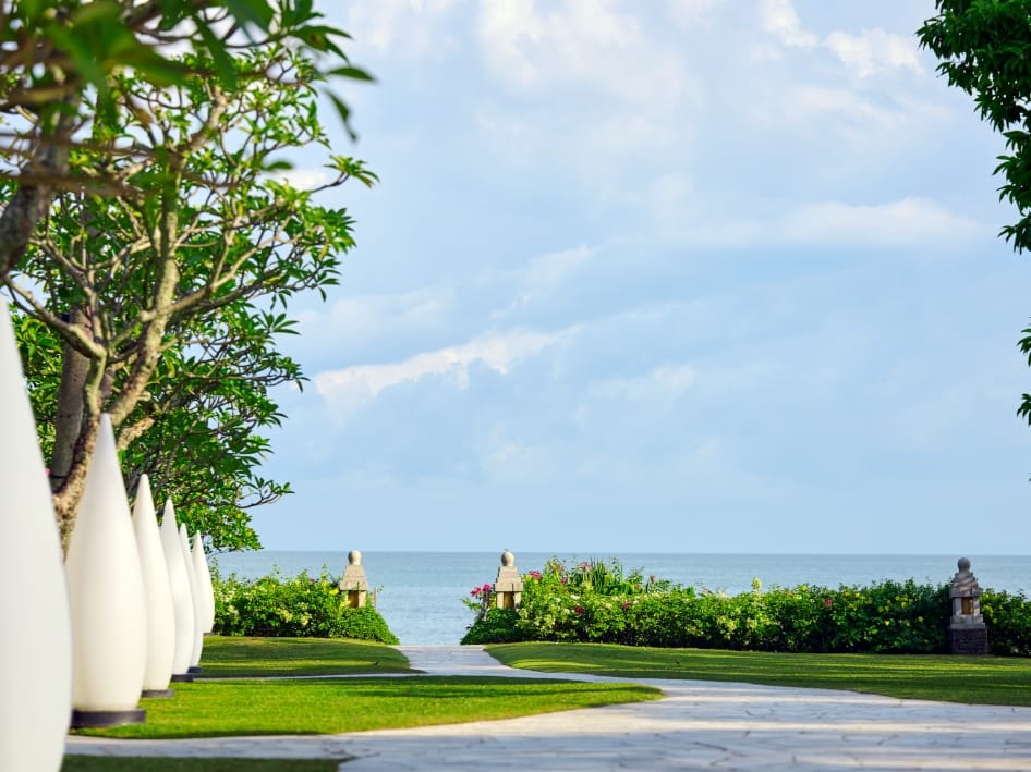 Path leading to the sea, flanked by decorative white cone sculptures and lush greenery while a paved walkway curves forward.