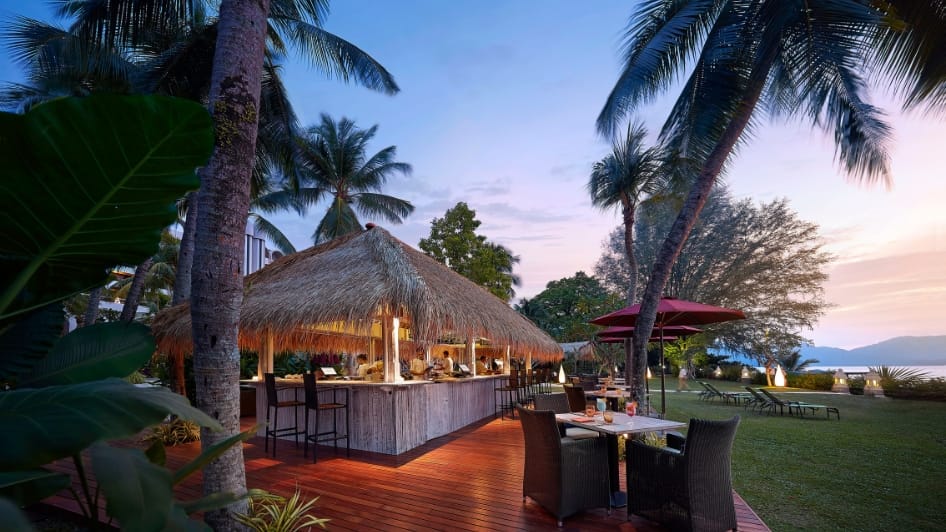 Tiki bar on a wooden deck at sunset, surrounded by palm trees and outdoor seating overlooking the water.